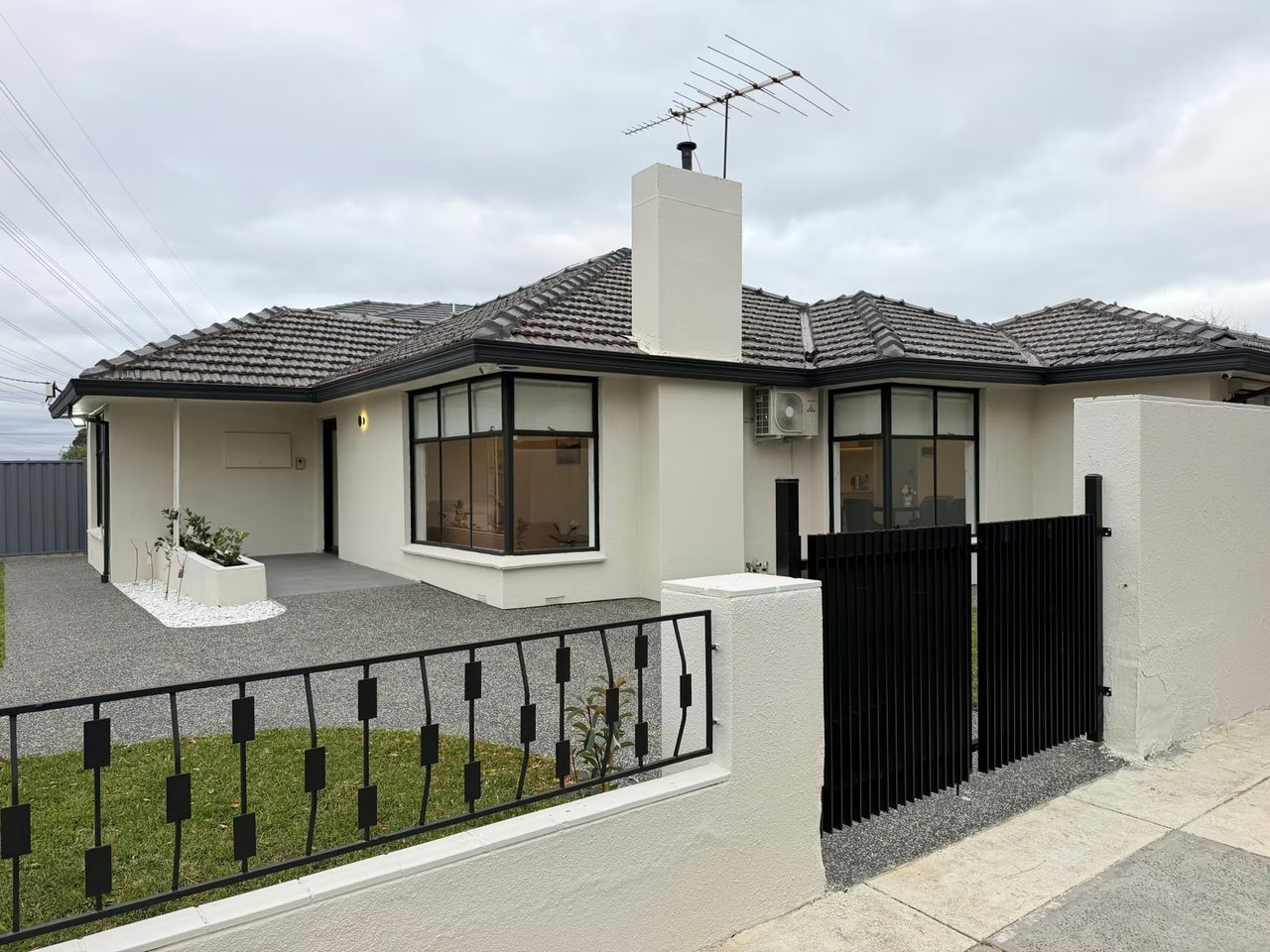 Street view of the single-storey Keilor East home — cream rendered walls, a dark tiled roof and black-framed windows — set behind a black steel fence and sliding gate, with a level exposed-aggregate forecourt running to the covered porch on the left.