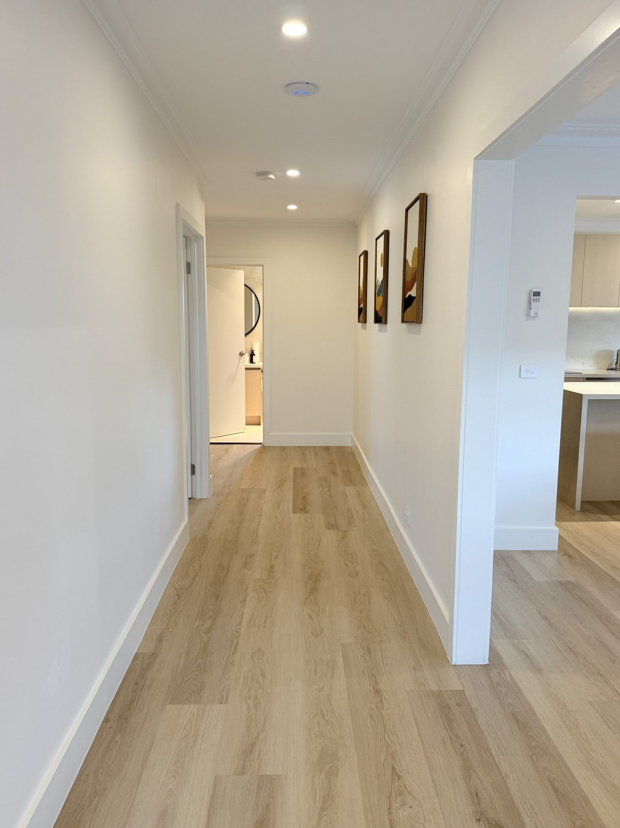A wide, step-free hallway in timber-look vinyl plank, running past a bathroom doorway at the far end and opening on the right into the kitchen. White walls carry three framed prints, with downlights and a smoke alarm set into the ceiling.