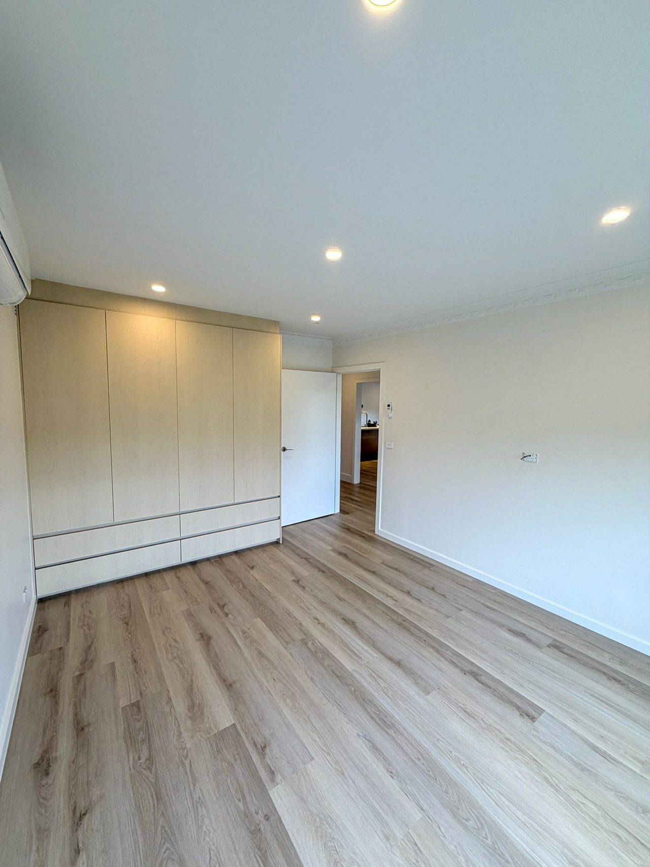 Bedroom one, with a full-width built-in wardrobe of four doors over deep drawers, a wall-mounted split-system air conditioner, and clear floor space between the wardrobe and the wide doorway out to the hall.