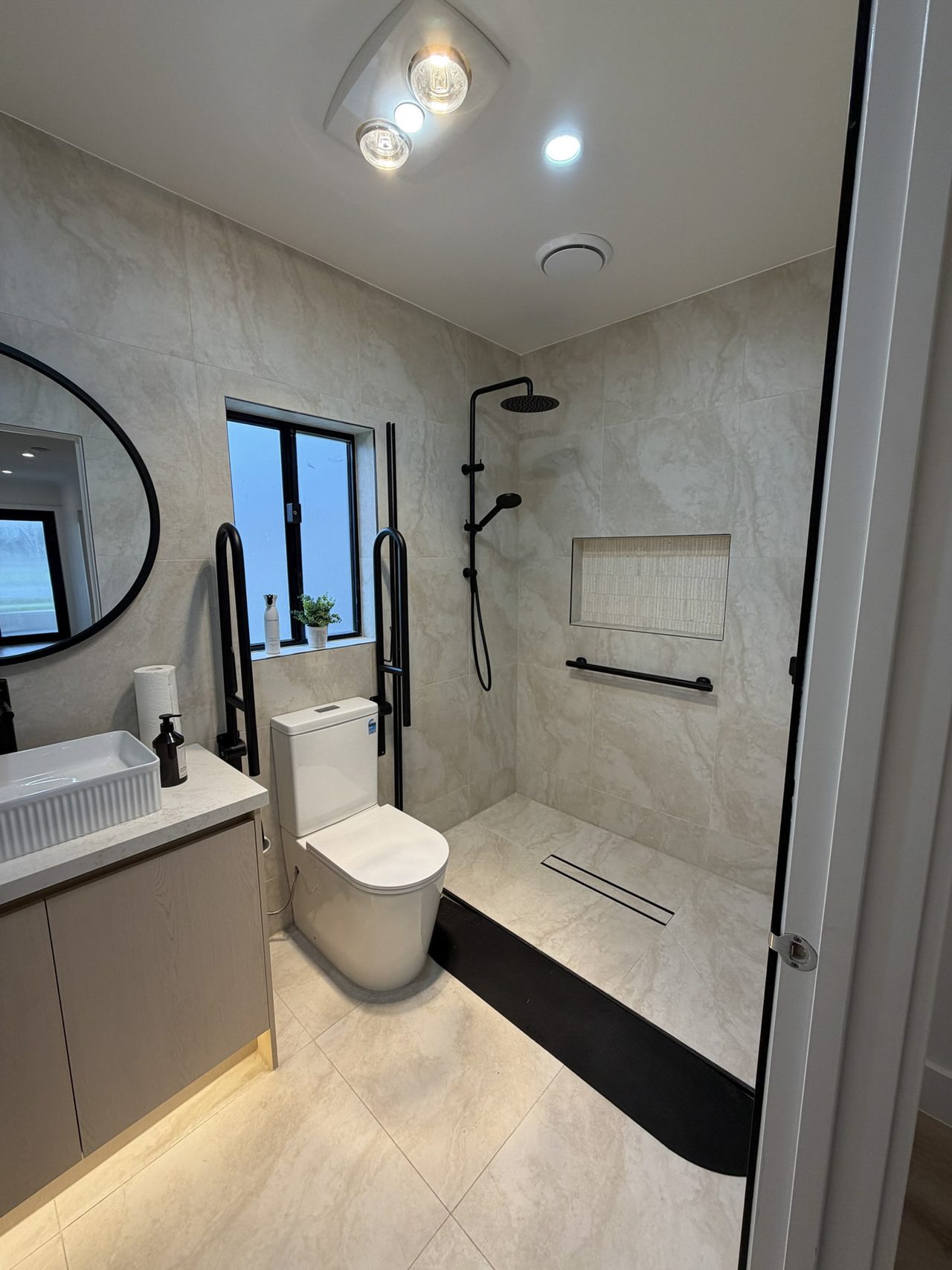The modified main bathroom: a step-free shower with no hob, a linear floor drain set flush into the tiles, a horizontal grab rail on the shower wall, and paired vertical grab rails beside the toilet. Both a rain head and a handheld shower on a slide rail are fitted, alongside a recessed wall niche, a vanity with basin, and a round mirror.