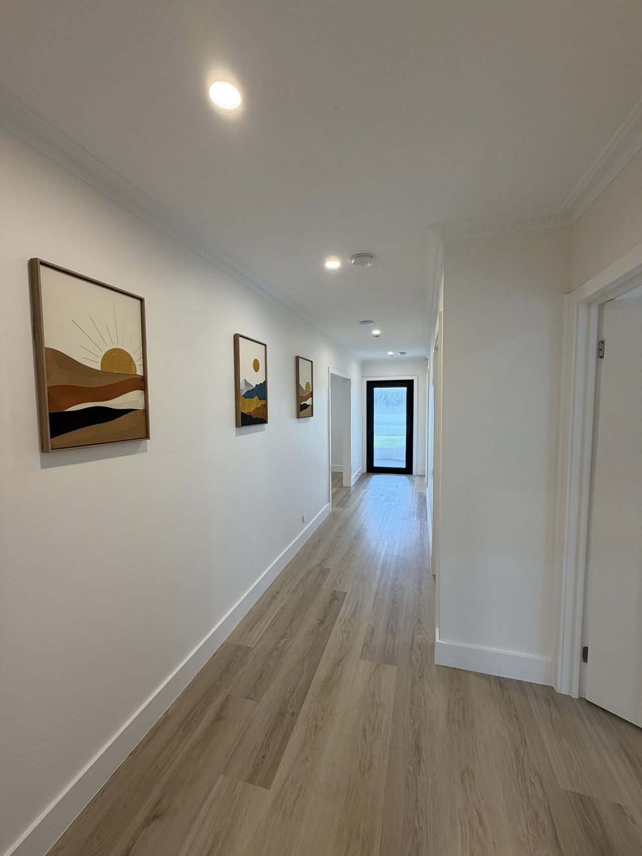 The hallway looking back from the bathroom towards the glazed front door — a long, unobstructed step-free run in vinyl plank, with framed prints along the wall and doorways opening off both sides.