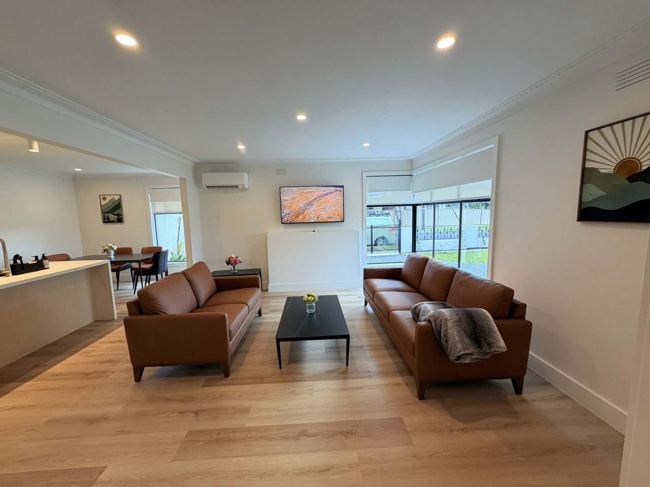 The open-plan living area: two tan leather sofas either side of a low black coffee table, a wall-mounted television, and large windows onto the front garden. The floor continues without a step into the kitchen and dining space beyond, with wide clear space between the furniture.