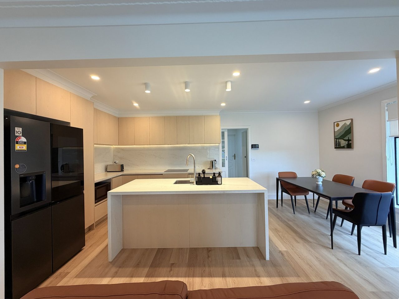 The kitchen and dining area: a large stone-topped island with an undermount sink, pale timber cabinetry with an under-lit stone splashback, a black French-door fridge, and a wall oven. A six-seat dining table sits alongside, with wide circulation space around the island.