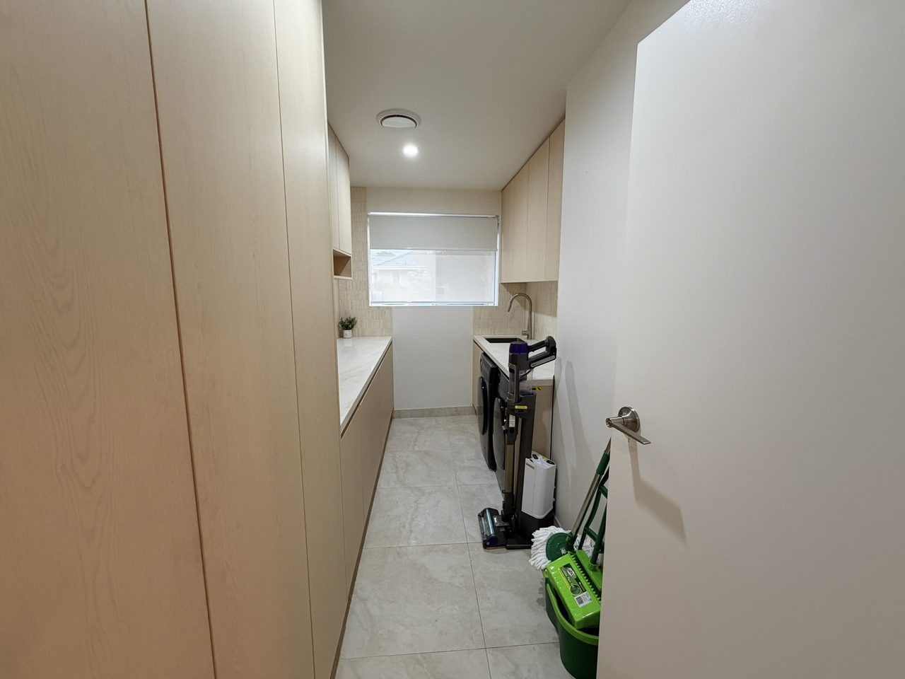 The internal laundry — a run of pale joinery under a stone benchtop along one wall, overhead cabinets, a sink and a front-loading washing machine, on tiled flooring. A window with a roller blind lights the far end.